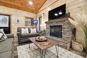 A cozy living room with a stone fireplace, a flat-screen TV, a sofa, and a coffee table with a plant and books. - Cozy Cardinal with Hot Tub (Sevierville)