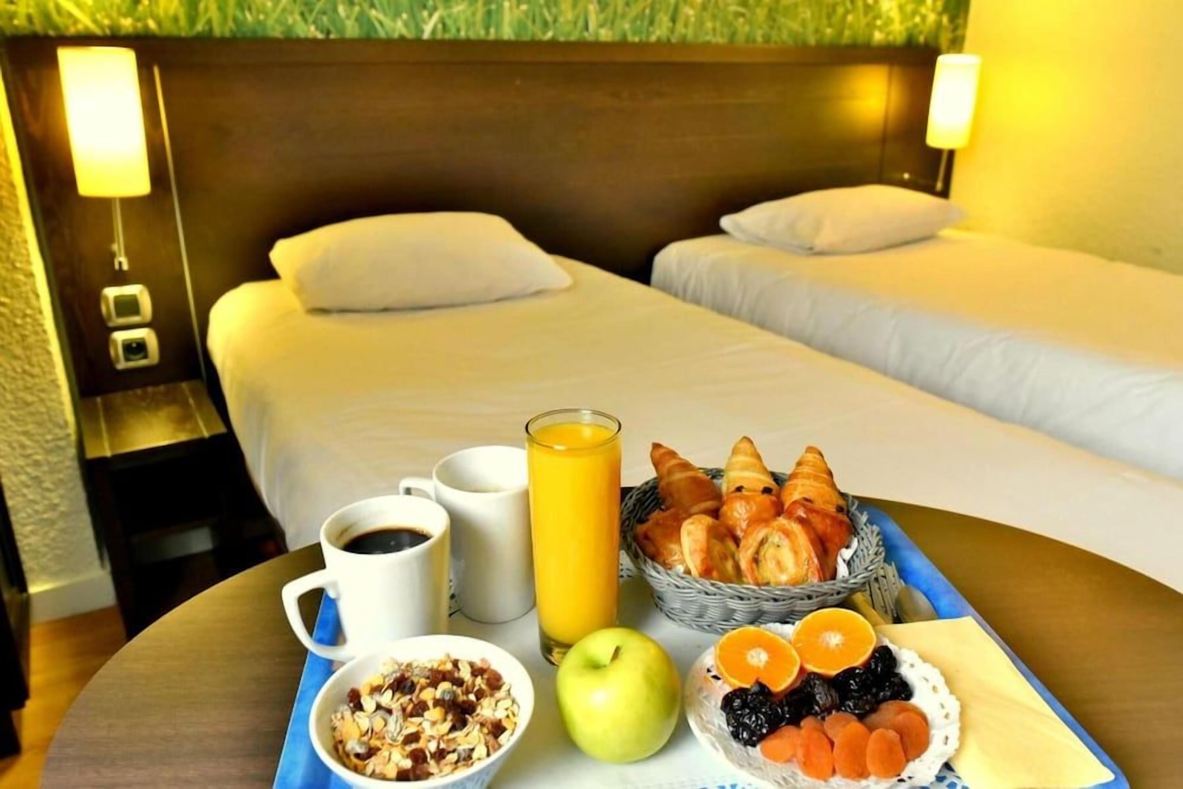 A hotel room with two beds, a breakfast tray with croissants, fruits, and a glass of orange juice.