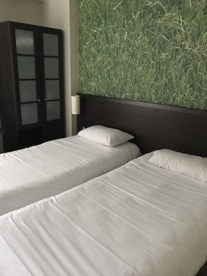 A hotel room with two single beds, a dark wooden wardrobe with frosted glass doors, and a wall with a green grass-like textured panel.