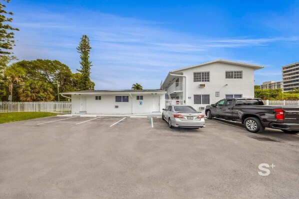 Un bâtiment à deux étages, à l’extérieur blanc, avec un parking où sont garées deux voitures et une clôture blanche qui entoure la propriété.