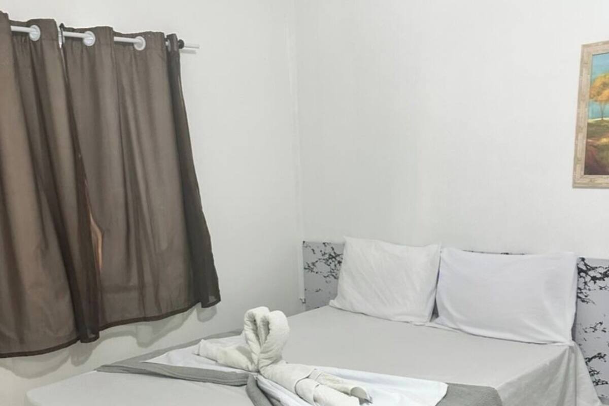 Una cama individual con un edredón gris, almohadas blancas y una toalla sobre la cama. Hay una mesita con mantel blanco y una toalla encima. La habitación tiene paredes blancas y una ventana con cortinas marrones.