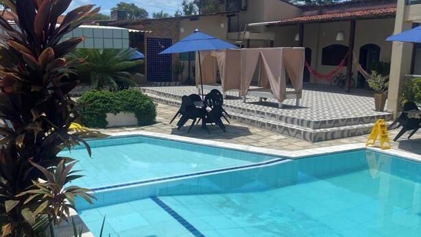 A swimming pool with a tiled deck, surrounded by plants and umbrellas. - Novo Hotel Maracaipe (Ipojuca)