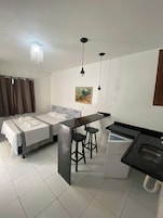 Quarto de hotel compacto com cama, pequeno espaço de cozinha, área de refeições com banquetas e um quadro na parede.