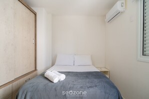 Ein kleines, ordentlich gemachtes Bett mit grauer Tagesdecke und zwei Kissen, ein Holzschrank, ein kleiner Nachttisch und eine Wand-Klimaanlage.