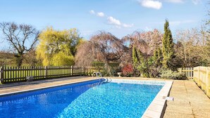 Una piscina rectangular con borde de azulejos, rodeada por una valla de madera y árboles con follaje otoñal al fondo.