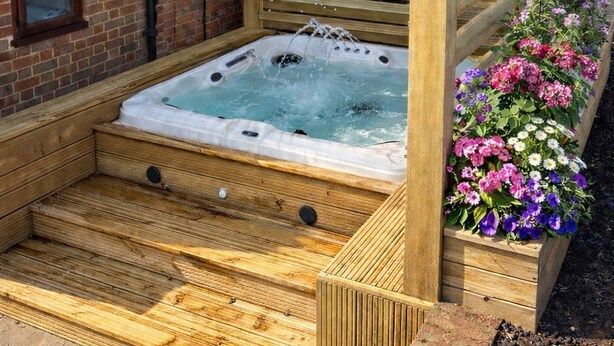 Una bañera de hidromasaje de madera con una pérgola de madera encima, rodeada de un muro de ladrillo y un parterre de flores.