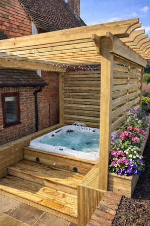 Una bañera de hidromasaje de madera con una pérgola de madera encima, rodeada de un muro de ladrillo y un parterre de flores.