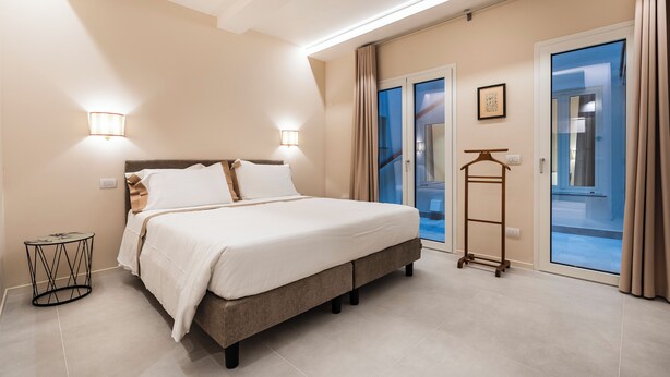 A bedroom with a bed, bedside lamps, a small round table, a coat rack, and a view through a glass door. - Palazzo Accademia-Brunelleschi-1 (Firenze)
