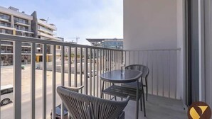 Un balcon avec une table et des chaises, donnant sur une rue bordée d’immeubles et de voitures garées.