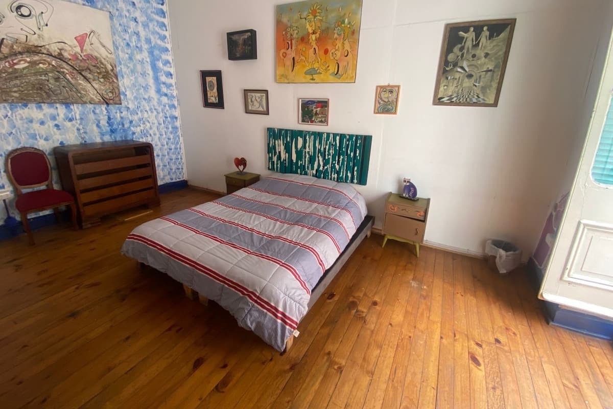 Un dormitorio con suelo de madera, una cama con colcha a rayas, una silla, un aparador y varias obras de arte en la pared.