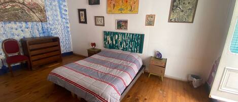 Un dormitorio con suelo de madera, una cama con colcha a rayas, una silla, un aparador y varias obras de arte en la pared.