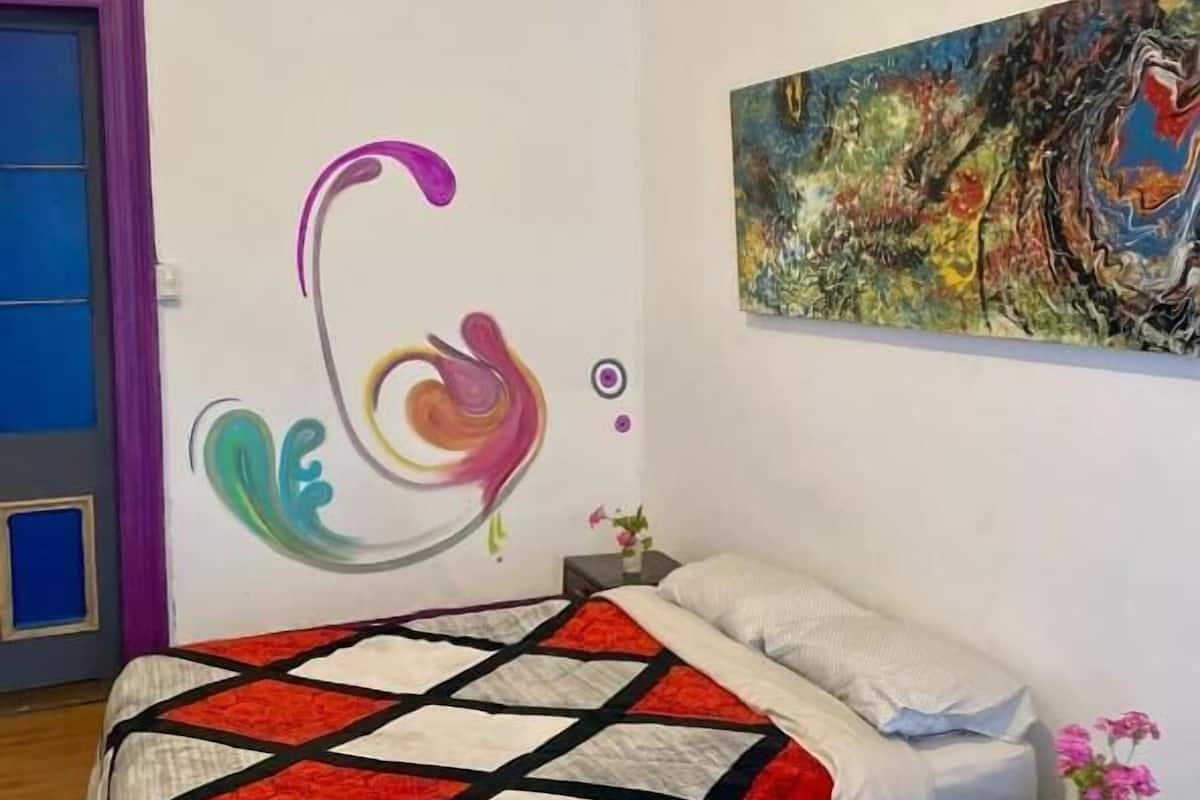 Un dormitorio con cama, un cuadro abstracto colorido y una puerta con cortinas moradas.