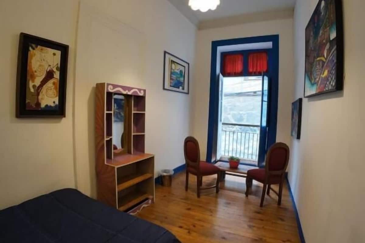 Una habitación con cama, dos sillas, una mesita y un ventanal con cortinas rojas.