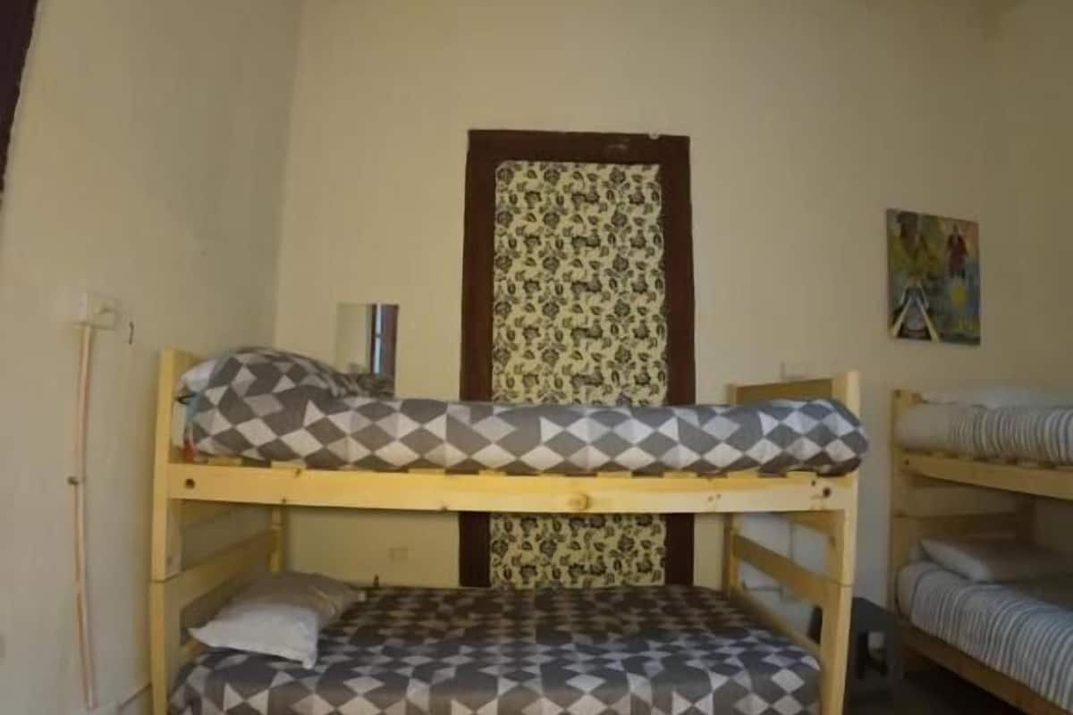 Habitación de residencia estudiantil con dos literas, un taburete pequeño y una puerta con cortina estampada.