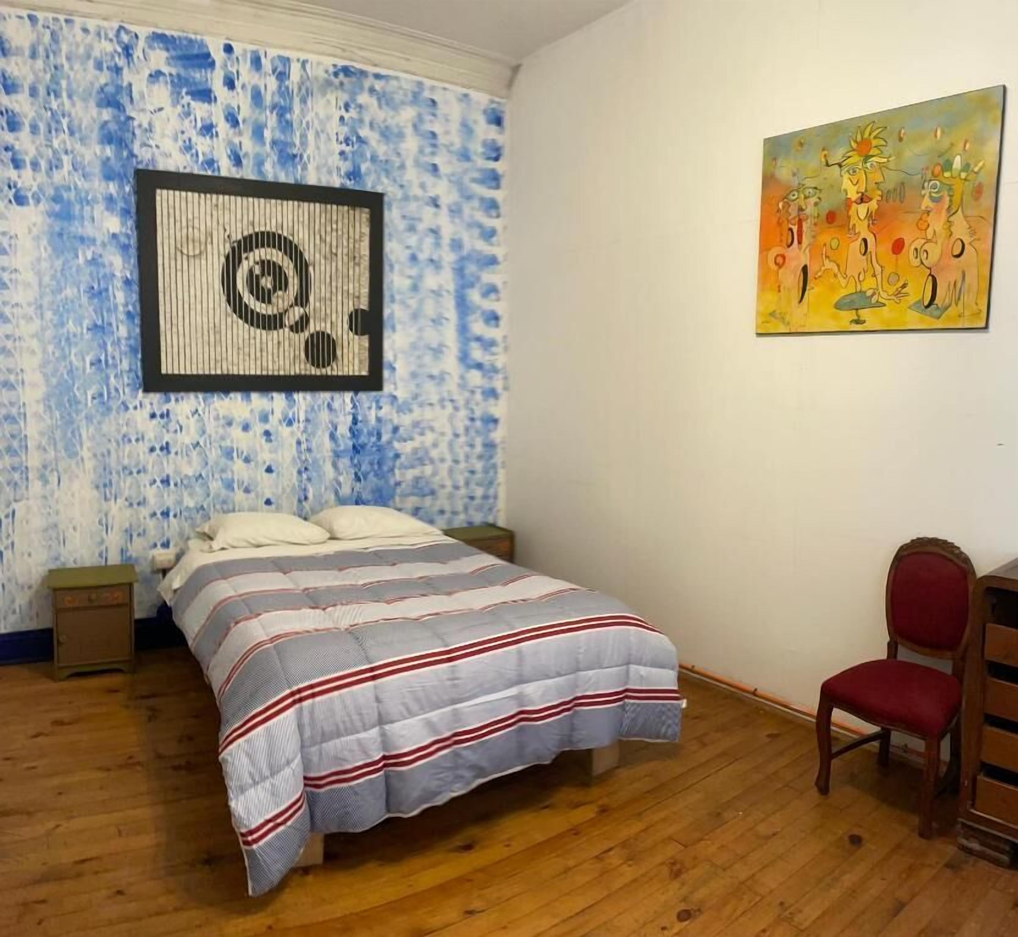 A bedroom with a bed, a nightstand, a chair, a wall art piece, and a wooden floor.