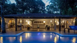 A poolside area with a covered seating area, a pool, and a reception desk. - 142D Casa com 03 Suítes em Trancoso (Porto Seguro)