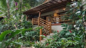 A wooden staircase leading to a covered area with a view of lush greenery and tropical plants. - 142D Casa com 03 Suítes em Trancoso (Porto Seguro)