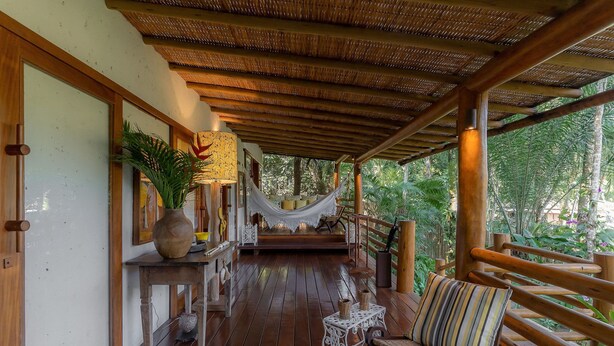 A wooden deck with a hammock, a bench, and a table with a vase and a lamp. - 142D Casa com 03 Suítes em Trancoso (Porto Seguro)