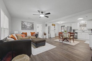 A spacious living room with a dark-colored sofa, a dining area with a wooden table and chairs, and a kitchen with white cabinets and appliances. - FriscoDowntown UniversalToyota Soccer3BDFullHome (Frisco)