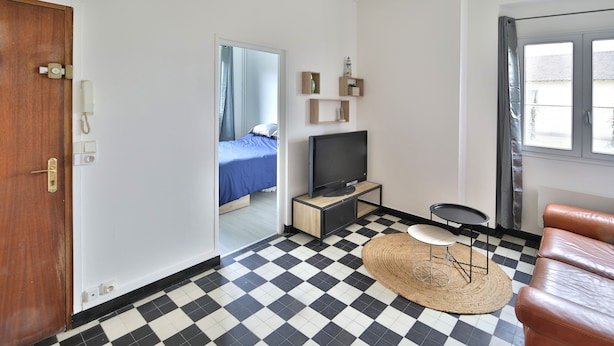 Una habitación con piso a cuadros, un televisor sobre un mueble, una pequeña mesa redonda, un sofá de cuero marrón y una puerta que da a otra habitación.
