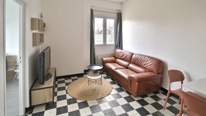 Una sala de estar con piso a cuadros blanco y negro, un sofá de cuero marrón, un televisor sobre un mueble de madera y una mesita redonda con camino de mesa tejido.