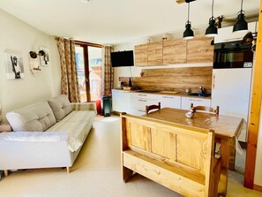 Un espace de vie compact comprenant un canapé, une table à manger en bois, un coin cuisine et une grande fenêtre.