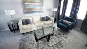 A living room with a beige sofa, a teal armchair, a glass coffee table, and two side tables with lamps. - Cute 4 Bed house - Central Tulsa with Fenced back yard (Tulsa)