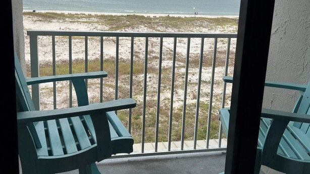 A balcony with two green chairs and a table, overlooking a beach with a sandy shore and a cloudy sky. - Cozy Corner Studio Unit - Perfect for 2! (Gulf Shores)