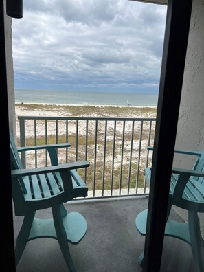 A balcony with two green chairs and a table, overlooking a beach with a sandy shore and a cloudy sky. - Cozy Corner Studio Unit - Perfect for 2! (Gulf Shores)