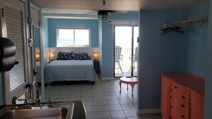 A hotel room with a bed, a bathroom with a sink, a balcony with chairs, and a view of the sea. - Cozy Corner Studio Unit - Perfect for 2! (Gulf Shores)