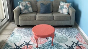 A living room with a grey sofa, three cushions, a red table, and a patterned rug. - Cozy Corner Studio Unit - Perfect for 2! (Gulf Shores)