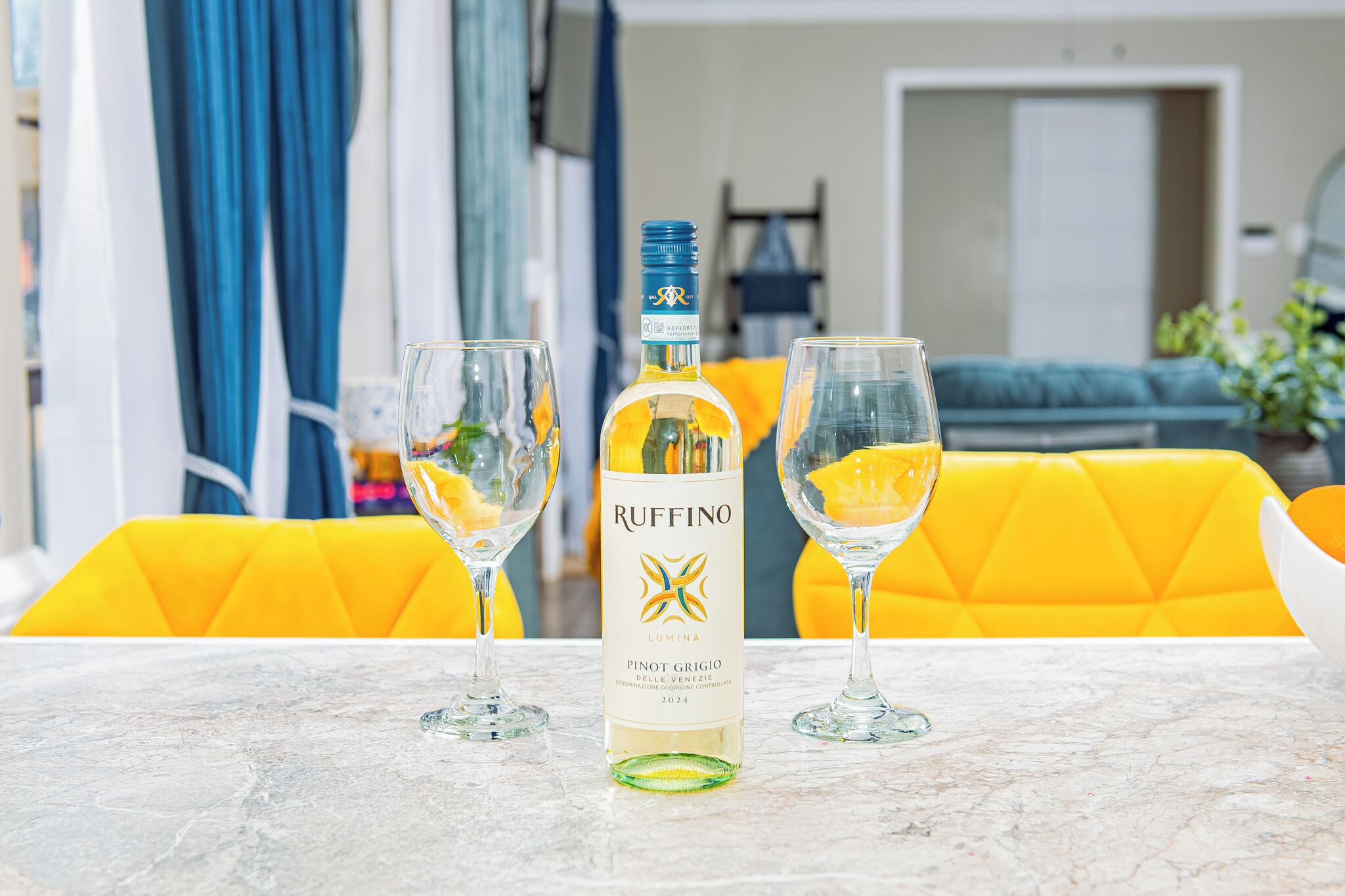 A bottle of Ruffino wine on a marble table with two glasses, a yellow sofa, and a blue curtain in the background.
