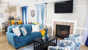 Ein Wohnzimmer mit einem blauen Sofa, einem Couchtisch und gemusterten Sesseln. Im Hintergrund ist ein Kamin zu sehen.