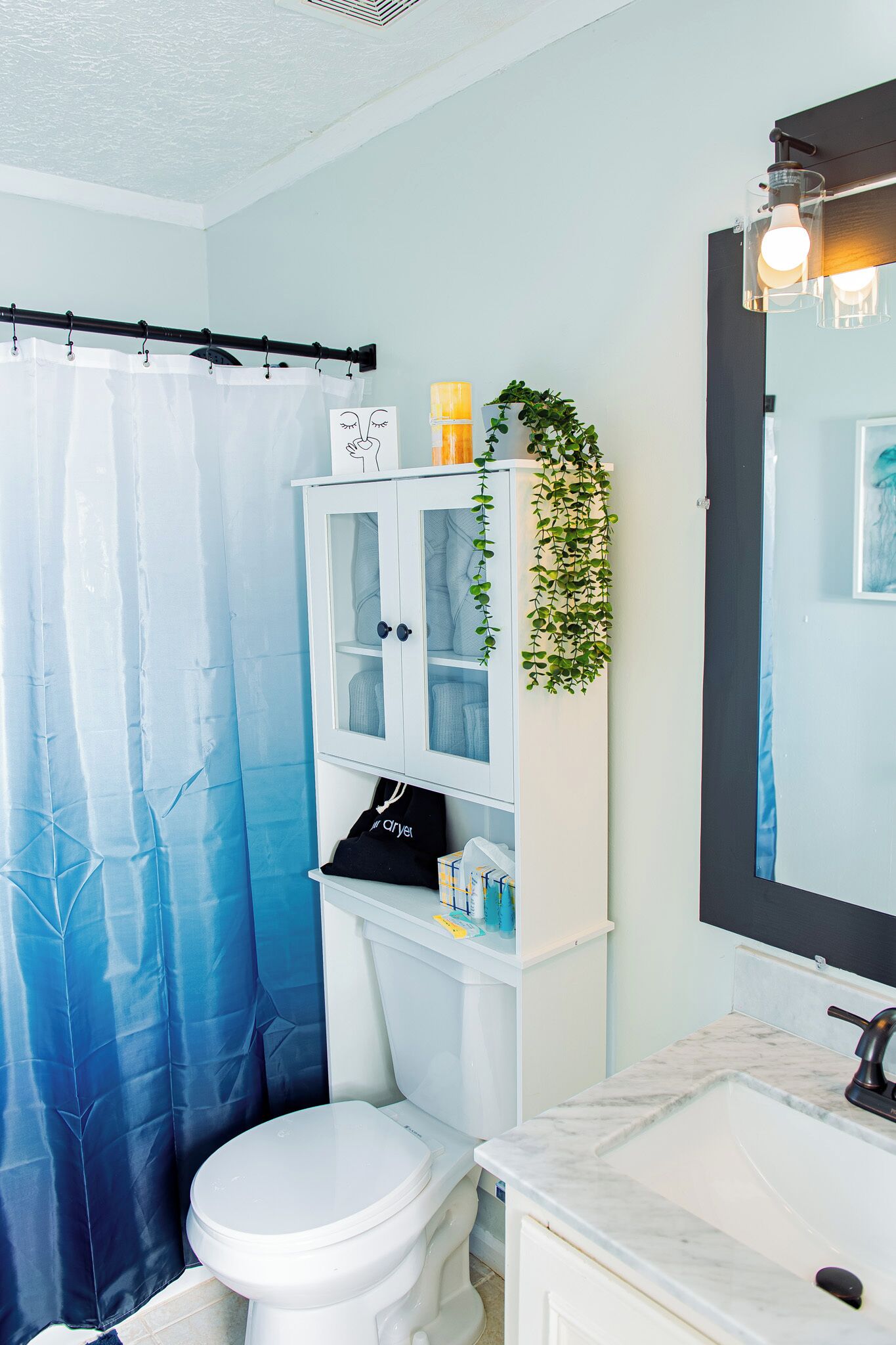Een badkamer met een wit toilet, een witte kast met glazen deuren, een blauw douchegordijn en een spiegel met verlichting.