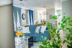 Un salón con un sofá azul, una mesita de centro y un tapete estampado. Se ve una ventana con cortinas azules y una planta en primer plano.