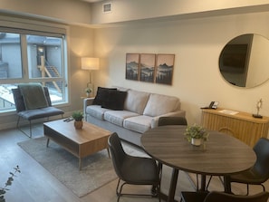 A modern living room with a grey sofa, a wooden coffee table, a round dining table with four chairs, a side table with a lamp, and a large window with a view of a staircase and a building outside. - Stylish, Modern One Bedroom Suite on the Second Level. Pool, Gym and Hot Tub! (Dead Man's Flats)