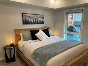 A bedroom with a wooden bed, a nightstand with a lamp, a framed picture on the wall, and a window with a view of the outside. - Stylish, Modern One Bedroom Suite on the Second Level. Pool, Gym and Hot Tub! (Dead Man's Flats)