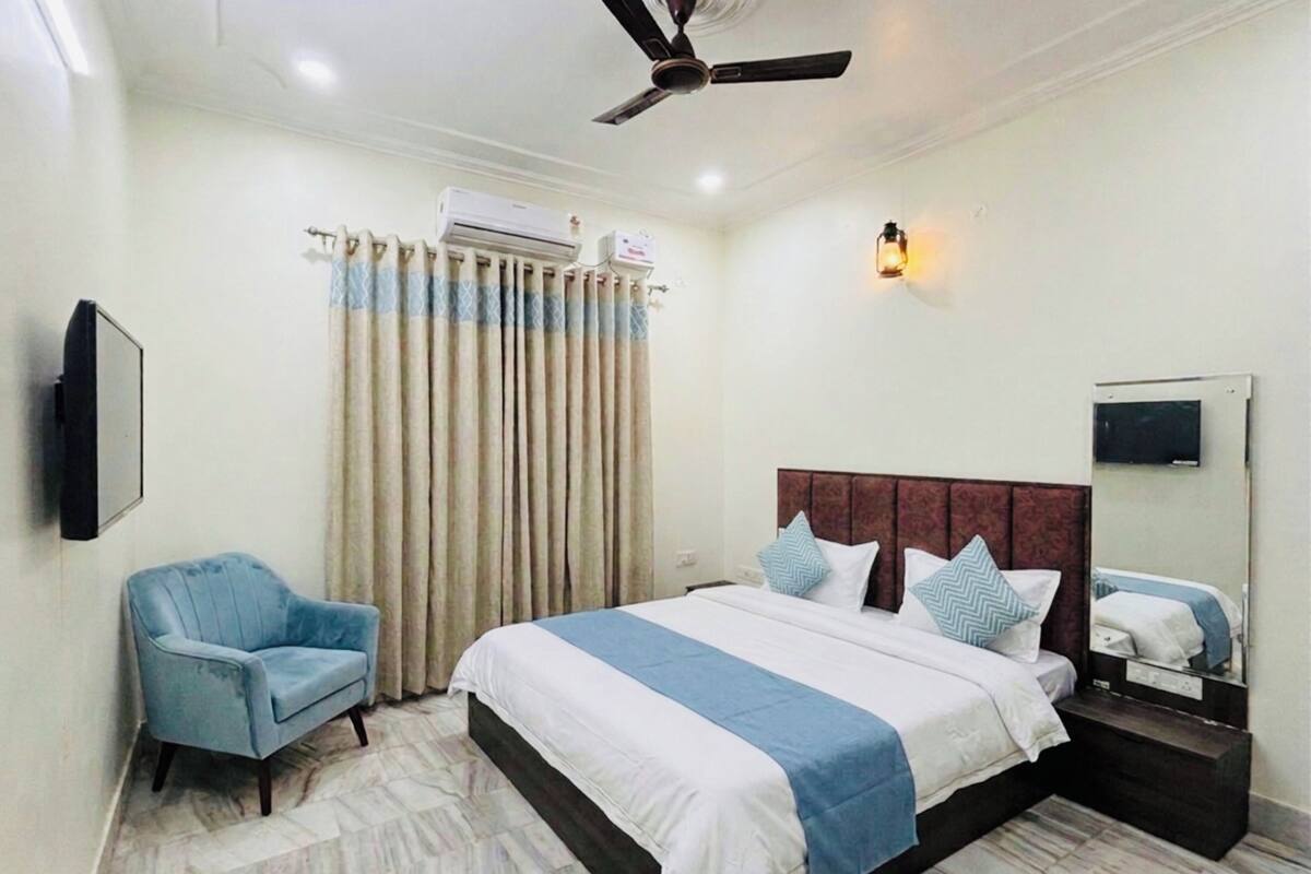 A hotel room with a bed, a chair, a ceiling fan, an air conditioning unit, a wall-mounted TV, and a nightstand.