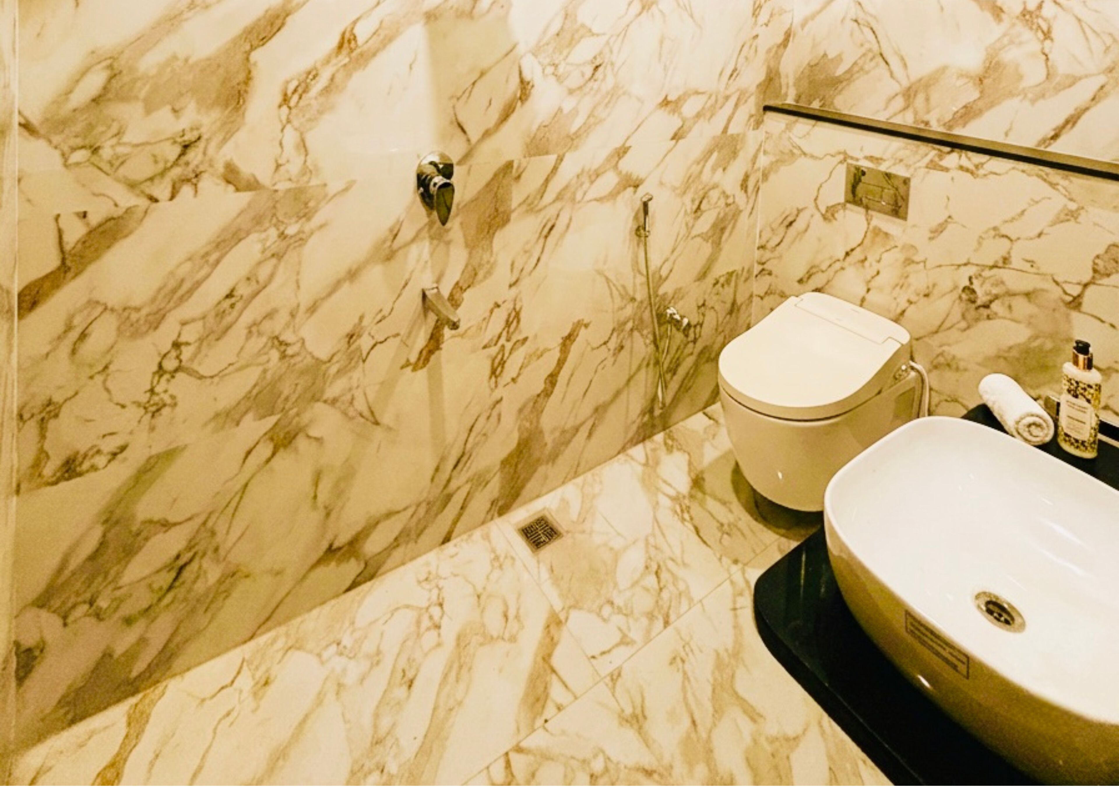 A bathroom with a marble wall, a toilet, a sink, and a towel dispenser.