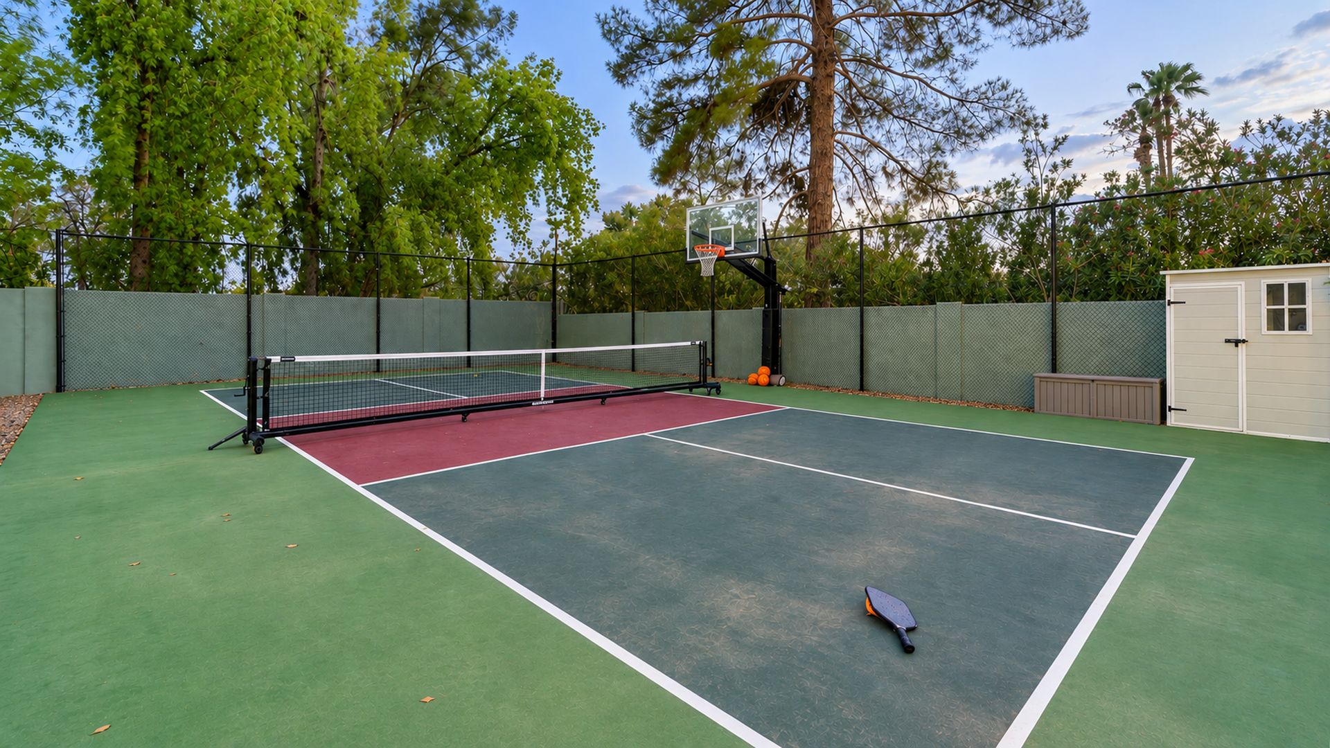 A tennis court with a net, surrounded by a fence, trees, and a shed.