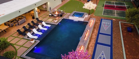 Una piscina con sillas de descanso y una chimenea, rodeada por una cancha de tenis y un área de juego de shuffleboard.