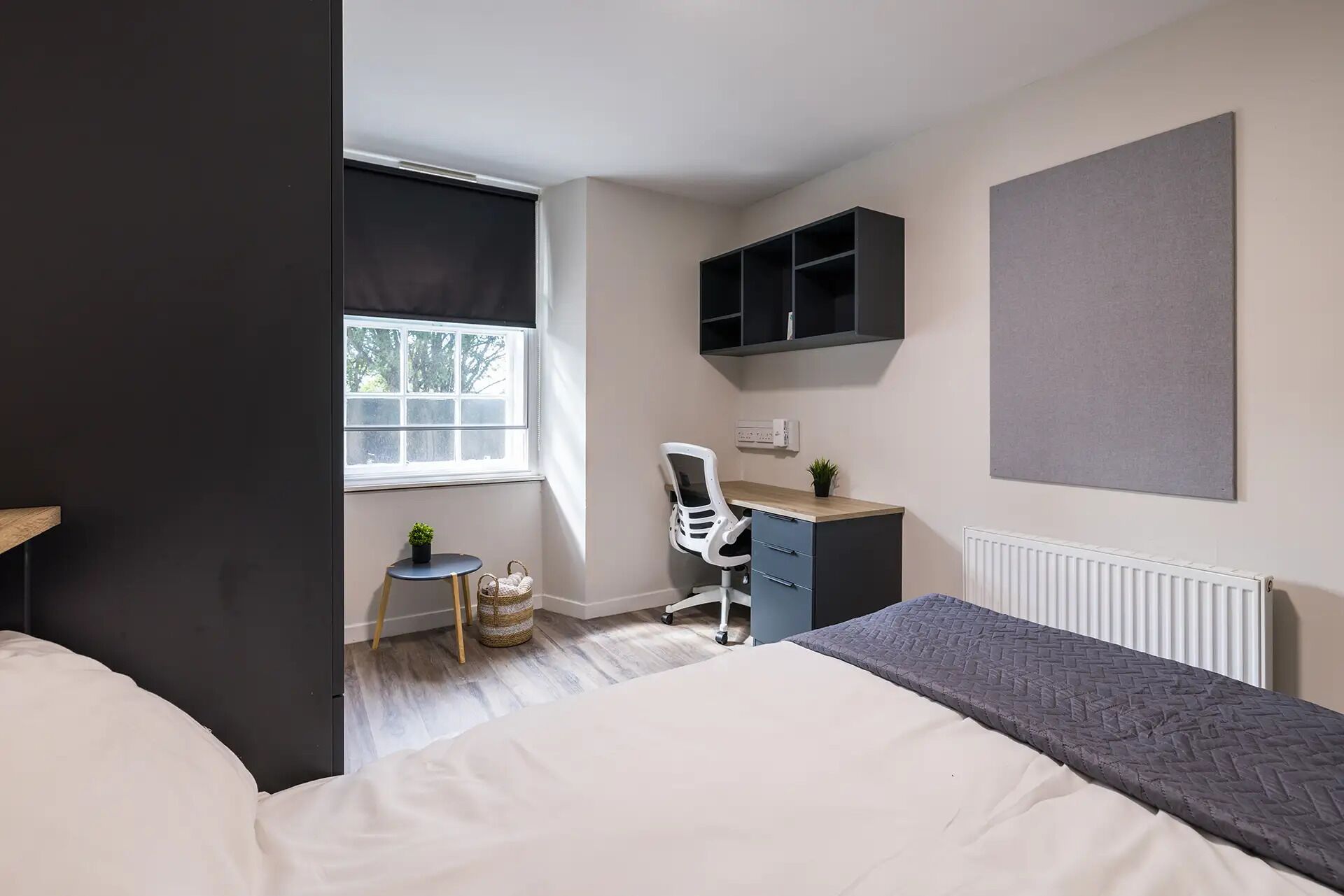 A bedroom with a bed, a desk with a chair, a radiator, a window, and a shelf.