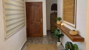 Un couloir avec une porte en bois, un luminaire au plafond, un tapis tissé et une étagère ornée d’objets décoratifs.
