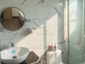 A bathroom with a round mirror, a white sink, a glass shower enclosure, and a marble wall. - Central Garden (Ho Chi Minh City)