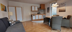 Un espacio habitable compacto con kitchenette, comedor con mobiliario de mimbre y sofá con cojín decorativo.