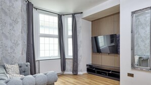 Een moderne woonkamer met een grijze bank, een flatscreen televisie bevestigd aan een houten paneel, en een groot raam met grijze gordijnen.