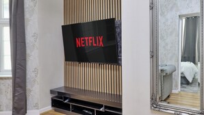 Een kamer met een groot flatscreen televisietoestel bevestigd aan een wand van houten panelen, een spiegel met een sierlijk frame en een bed met een grijze hoofdbord.