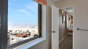 A hotel room with a large window overlooking a beach, a hallway leading to a bathroom, and a bed with a nightstand. - Two Pina Coladas at San Luis Resort (Galveston)