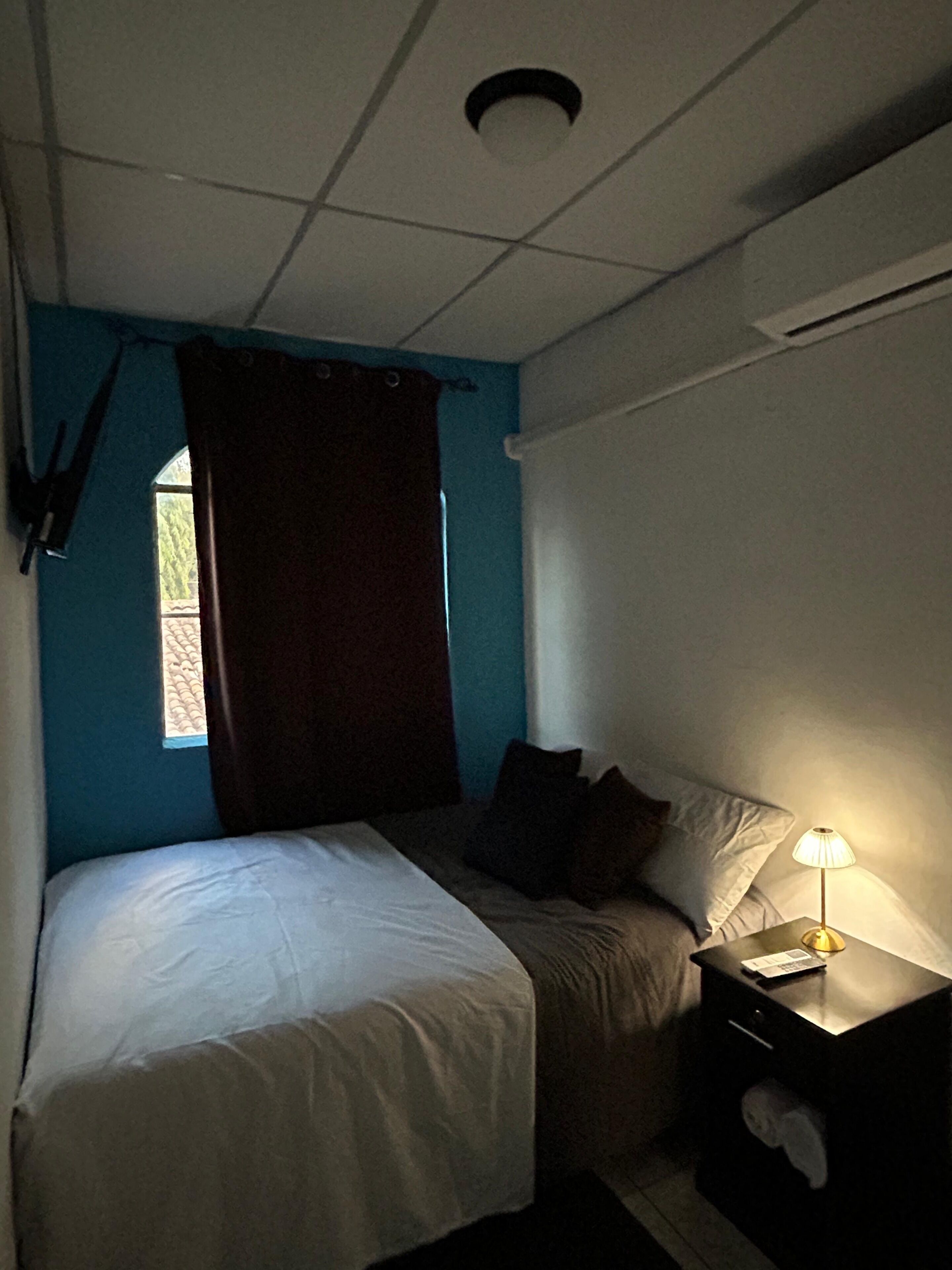 Una habitación pequeña y tenuemente iluminada con una cama individual, una mesita de noche con lámpara y una ventana con cortinas.