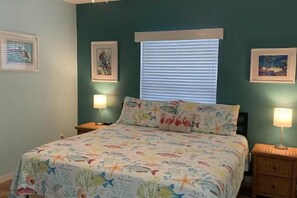 Un dormitorio con cama, dos mesitas de noche, una ventana con persianas y cuadros enmarcados en las paredes.